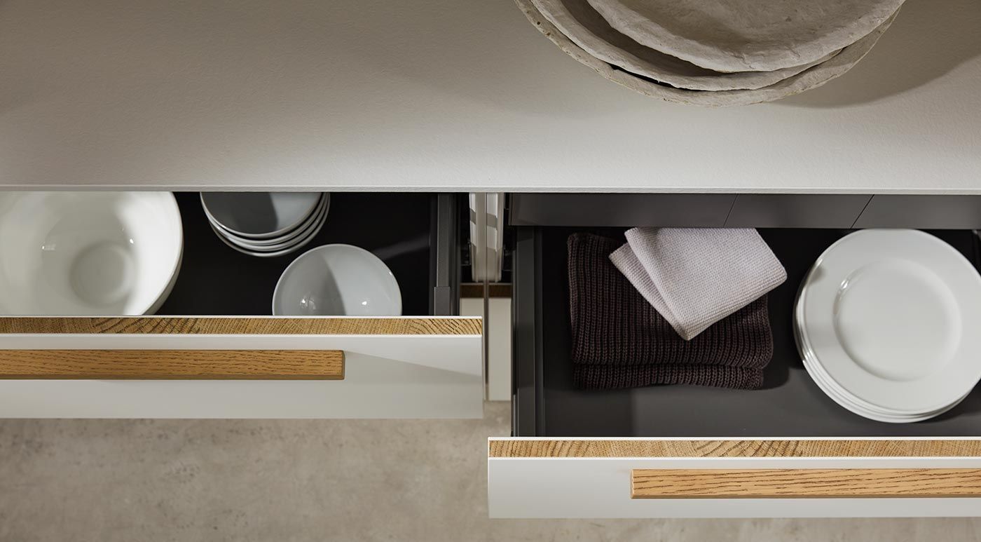 Geöffnete weiße Schubladen mit Geschirr, Textilien und markanten Eichenholz-Griffleisten.