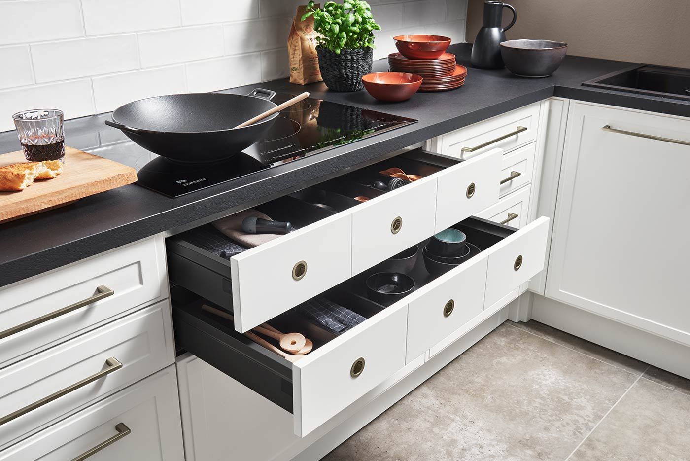 Geöffnete weiße Schubladen unter schwarzer Arbeitsplatte mit Wok und Geschirr.